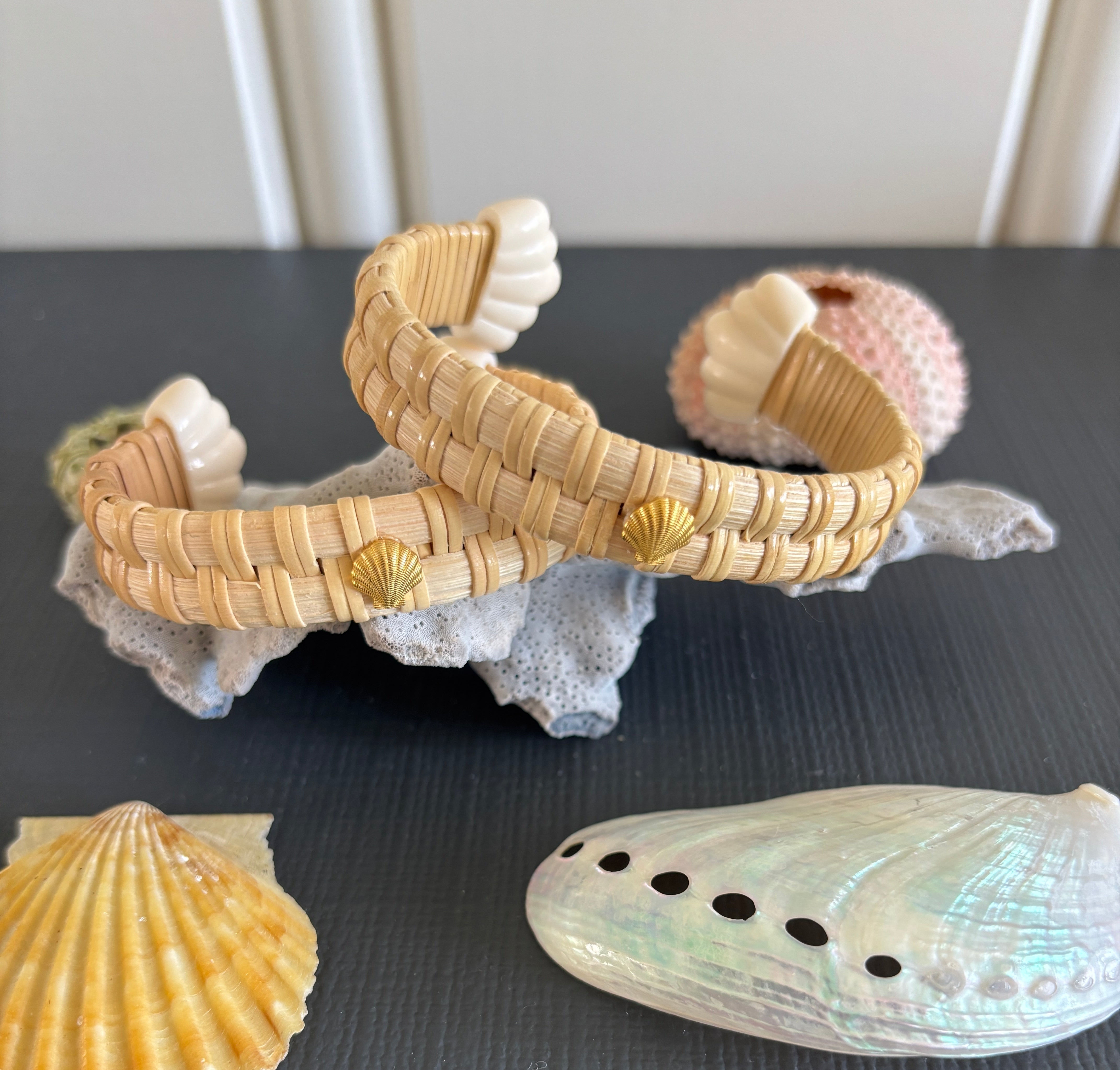Two woven nantucket basket cuff bracelets decorated with small seashells, displayed on a white decorative rock alongside various real seashells on a dark gray surface.